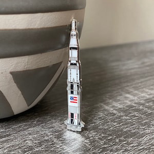 Può includere: Uno spillo in smalto argentato a forma di razzo con una bandiera americana sul lato. Il razzo è in piedi su una superficie di legno grigia.