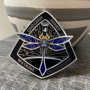 May include: A black enamel pin with a silver border, featuring a dragonfly with blue wings and a yellow body. The pin is shaped like a diamond and has the names "Lindgren", "Hines", "Watkins", and "Cristoforetti" around the edges.