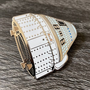 Op de afbeelding: Een gouden en witte emaille pin van een ruimtecapsule met een gedetailleerd ontwerp. De capsule is wit met gouden accenten en blauwe details.