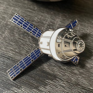 May include: A silver and white enamel pin of a space capsule with blue solar panels. The capsule is white with silver details and the solar panels are blue with silver outlines.