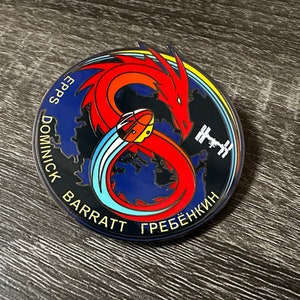 May include: A round, blue and black enamel pin with a red dragon wrapped around a blue and white comet. The pin features the text "EPPS DOMINICK BARRATT ГРЕБЁНКИН" around the edge.