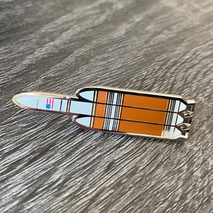 May include: A silver and orange enamel pin of a rocket with an American flag on the side.