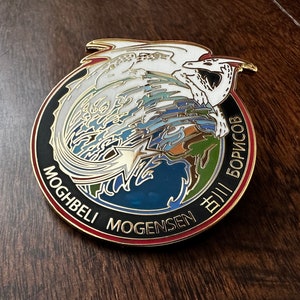 Op de afbeelding: Een gouden emaille pin met een witte draak die om een blauwe en groene wereld draait. De wereld heeft een gouden ster erop. De pin heeft een zwarte achtergrond met de tekst "MOGHBELI MOGENSEN ДМИТРИЙ БОРИСОВ" rond de rand.