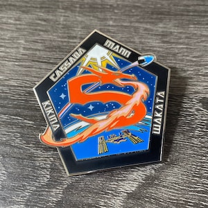 May include: A black and blue enamel pin with a red dragon design. The pin features the names "Cassada", "Mann", "Wakata", and "Kikuina" around the perimeter. The dragon is flying in front of a blue and white background with stars.