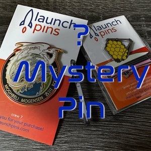 May include: Two enamel pins on cards. One pin features a dragon design with text, the other a telescope. The cards read "launch pins" and "Mystery Pin". The dragon pin has the text "MOGHBELI MOGENSEN 古川 50".