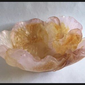 May include: A pink and gold glass bowl shaped like a flower. The bowl has a delicate, ruffled edge and a center with a raised, textured design.