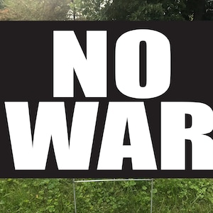 May include: A black rectangular sign with the words "NO WAR" in large, bold, white letters. The sign is displayed outdoors, mounted on a metal frame, and set against a backdrop of green grass and foliage.