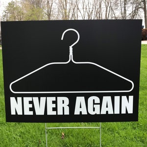 May include: A black yard sign with a white outline of a coat hanger and the text "NEVER AGAIN" in white.