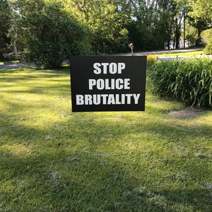 Pode incluir: Um sinal preto com texto branco que diz "STOP POLICE BRUTALITY" &eacute; exibido em um gramado verde.