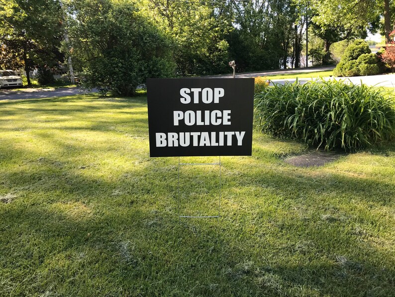 Pode incluir: Um sinal preto com texto branco que diz "STOP POLICE BRUTALITY" &eacute; exibido em um gramado verde.