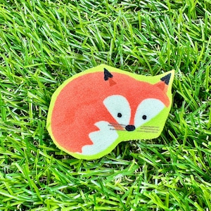 May include: A red and white fox applique with black eyes and a black nose. The fox is lying down with its head turned to the side. The applique is on a green grass background.