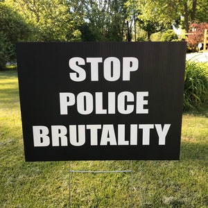 May include: A black sign with white text that reads "STOP POLICE BRUTALITY" on a green lawn.