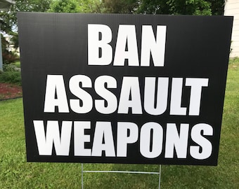 May include: Black and white sign with the text "BAN ASSAULT WEAPONS" in large, bold white letters.