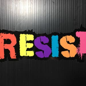 Puede incluir: Una pegatina con la palabra "RESIST" en colores arcoíris. Cada letra es de un color diferente, con un fondo negro y un aspecto texturizado y desgastado.