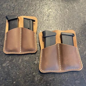 Może przedstawiać: Dwa brązowe skórzane etui na magazynki z białymi przeszyciami, każde mieszczące dwa czarne magazynki. Etui mają prostokątny kształt z zaokrąglonymi rogami i są umieszczone na ciemnym tle. Skóra ma naturalny, teksturowany wygląd.