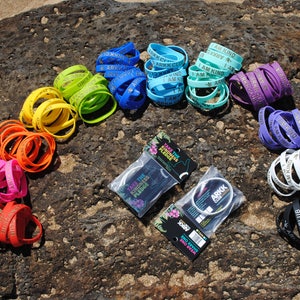 Puede incluir: Un surtido arcoíris de pulseras de silicona de colores con el texto "I AM KIND" y "ARKK CLUB" impreso en ellas. Las pulseras están dispuestas en forma de arcoíris sobre una superficie rocosa gris oscuro. Dos paquetes de pulseras son visibles en el centro inferior de la imagen. Los paquetes están etiquetados como "TAKE THE KINDNESS PLEDGE".