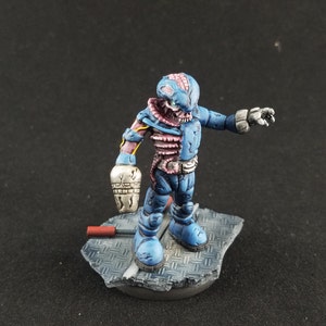 May include: A blue and pink painted miniature figure of a robot with a damaged body. The robot is standing on a gray base with a red and black object in the background.