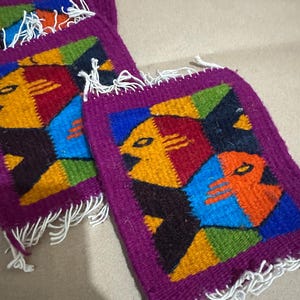 May include: Set of three square woven coasters. Each coaster features a colorful geometric design with a face-like motif in shades of yellow, red, blue, and green, framed by a purple border and white fringe.