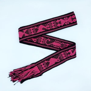 Pode incluir: Um cinto tecido preto e rosa com franja em uma extremidade. O cinto tem um padrão geométrico.