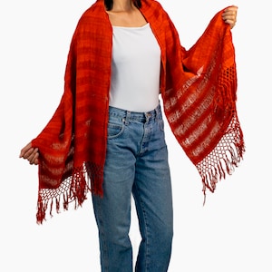May include: A woman wearing a red and white striped shawl with fringe. The shawl is made of a soft, woven fabric. She is also wearing a white t-shirt and blue jeans.