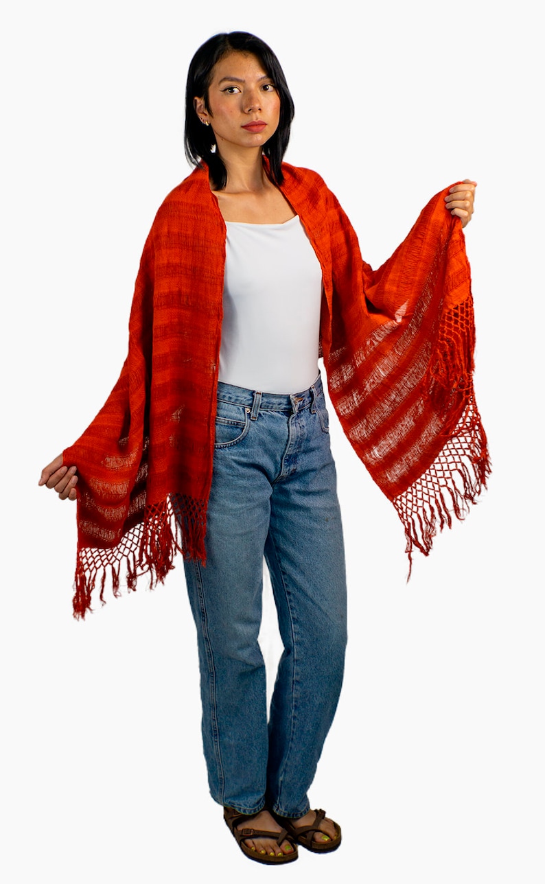 May include: A woman wearing a red and white striped shawl with fringe. The shawl is made of a soft, woven fabric. She is also wearing a white t-shirt and blue jeans.