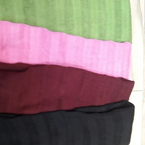 May include: Four woven scarves in different colors: green, pink, burgundy, and black. The scarves are stacked on top of each other.