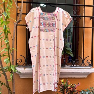 May include: A white cotton dress with colorful embroidery. The dress has a square neckline and short sleeves. The embroidery is in a geometric pattern and includes red, blue, green, yellow, and pink threads.