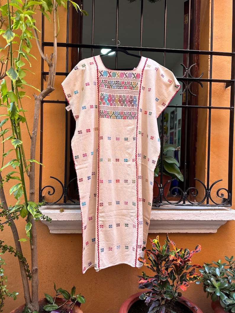 May include: A white cotton dress with colorful embroidery. The dress has a square neckline and short sleeves. The embroidery is in a geometric pattern and includes red, blue, green, yellow, and pink threads.