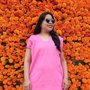 Op de afbeelding: Een vrouw in een roze geborduurde jurk en zonnebril staat voor een muur van oranje margrieten.