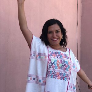 May include: A woman wearing a white embroidered blouse with colorful floral patterns. The blouse has a loose fit and a round neckline. The woman is smiling and has her arm raised in the air.