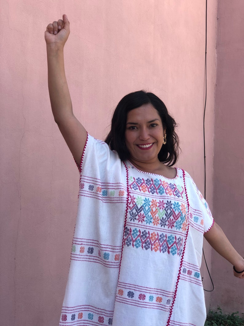 May include: A woman wearing a white embroidered blouse with colorful floral patterns. The blouse has a loose fit and a round neckline. The woman is smiling and has her arm raised in the air.