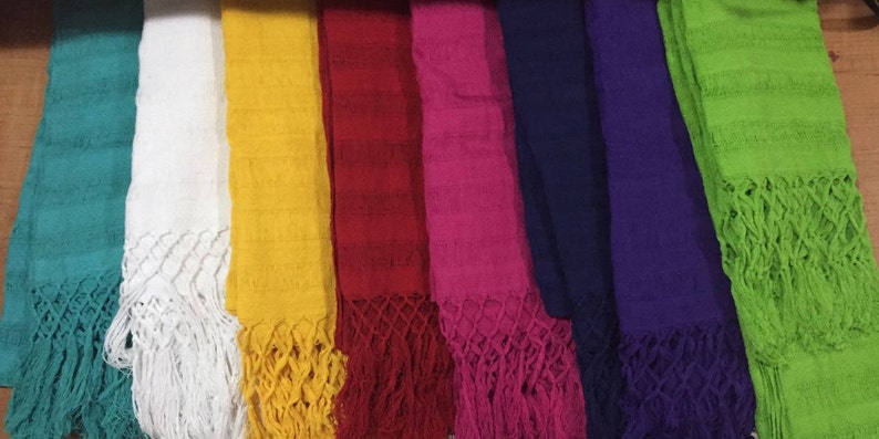 May include: A collection of seven colorful woven shawls with fringed edges. The shawls are arranged in a row, with the colors ranging from teal to green.