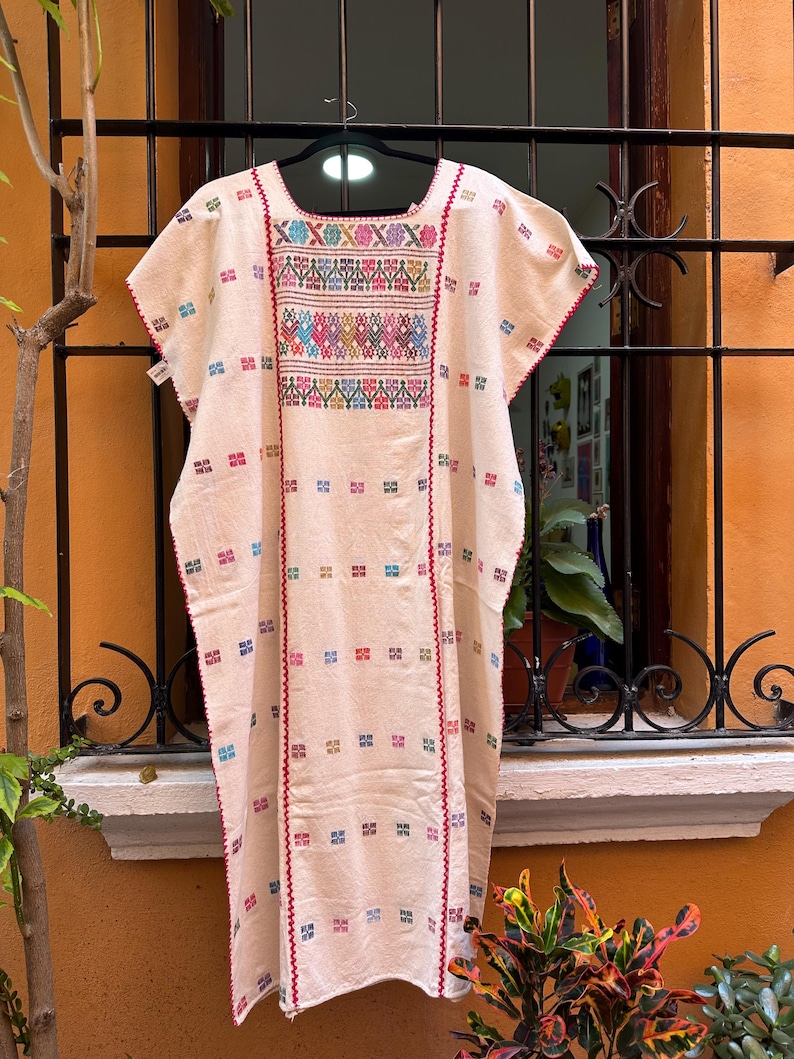 May include: A white cotton tunic with colorful embroidery. The embroidery features geometric patterns in shades of pink, blue, green, yellow, and red. The tunic has a scoop neck and short sleeves.