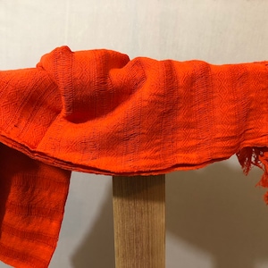 May include: A bright orange woven shawl with a fringe border. The shawl is folded and draped over a wooden surface.