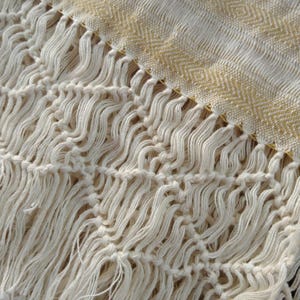 May include: Close-up of a cream-colored woven textile with a textured fringe. The fabric has a geometric pattern in a light gold color. The fringe is knotted and adds a decorative element to the textile.