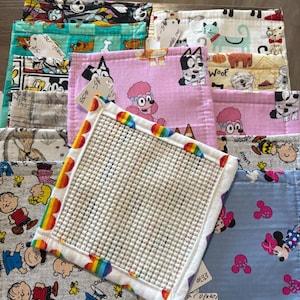May include: A stack of colorful fabric squares with various cartoon character prints, including Snoopy, Mickey Mouse, and Bluey. One square features a rainbow border and a textured surface. The fabrics are in shades of blue, pink, and green.