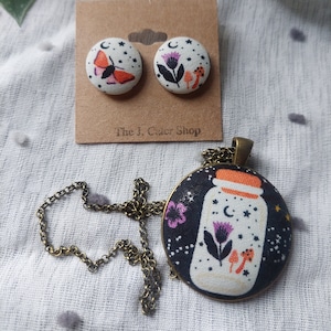 May include: A set of fabric earrings and a necklace featuring a white jar with a moon, stars, flowers, and mushrooms on a black background. The earrings and necklace are on a white fabric background.