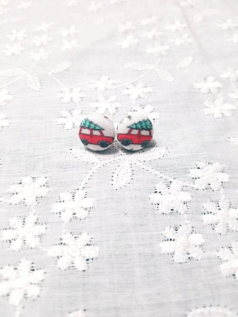 May include: A pair of red and green Christmas-themed fabric earrings with a car carrying a Christmas tree. The earrings are on a white lace background.
