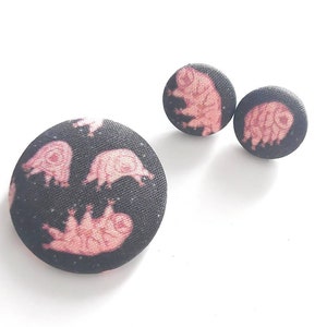 May include: A set of three black fabric buttons with a pink and white print of tardigrades. The buttons are round and have a diameter of approximately 1 inch.