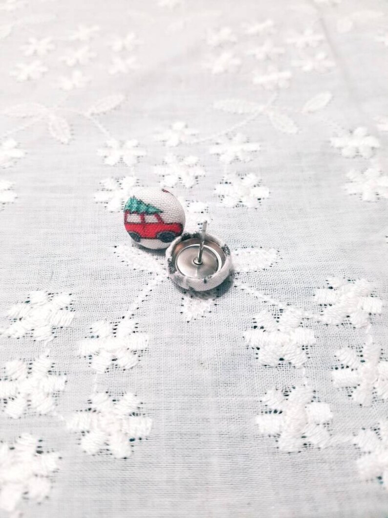 May include: A pair of fabric covered button earrings with a red car with a green Christmas tree on top. The buttons are white with a silver metal backing.