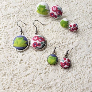 May include: A set of four fabric covered button earrings. Two pairs of earrings feature a green frog design, and the other two pairs feature a red and white floral design.