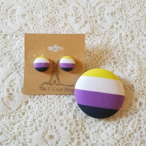 May include: A pair of non-binary pride flag earrings and a button pin. The earrings and pin are yellow, white, purple, and black.
