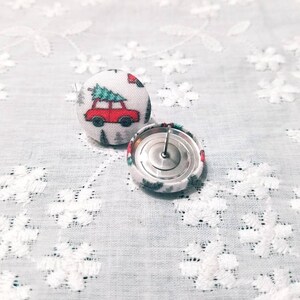 May include: A pair of Christmas-themed fabric button earrings. The buttons are white with a red car carrying a green Christmas tree. The back of one button is visible, showing a silver metal post.