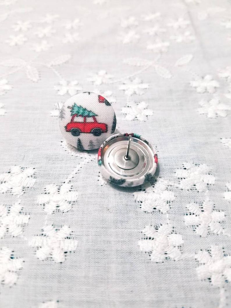 May include: A pair of Christmas-themed fabric button earrings. The buttons are white with a red car carrying a green Christmas tree. The back of one button is visible, showing a silver metal post.