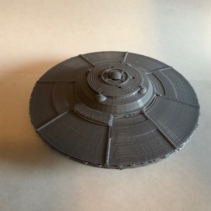 May include: A gray 3D printed model of a flying saucer. The saucer has a circular base with a raised center dome. The base has a series of concentric rings and radial lines.