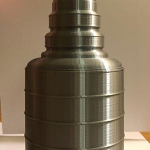 May include: A silver-colored 3D printed trophy replica featuring a wide bowl top with a textured rim, a tapered neck section, and a cylindrical base with horizontal ring details. The object has a visible layered finish from the printing process.