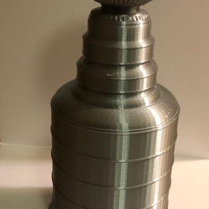 May include: A silver-colored trophy cup featuring a wide, bowl-shaped top with a textured rim, a narrow neck, and a multi-tiered cylindrical base. The object has a metallic finish with visible horizontal layer lines throughout its structure.