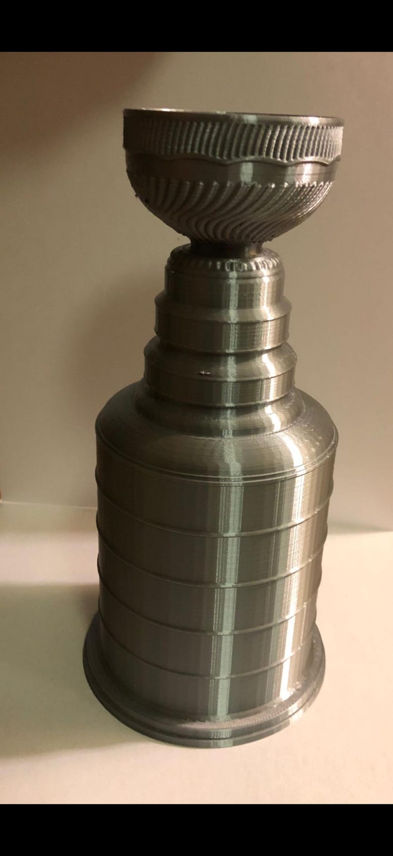May include: A silver-colored trophy cup featuring a wide, bowl-shaped top with a textured rim, a narrow neck, and a multi-tiered cylindrical base. The object has a metallic finish with visible horizontal layer lines throughout its structure.