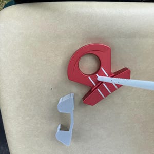 May include: A red golf putter with white stripes and a white shaft, along with a detached gray piece, rests on a beige surface. The putter has a circular opening and a unique design.