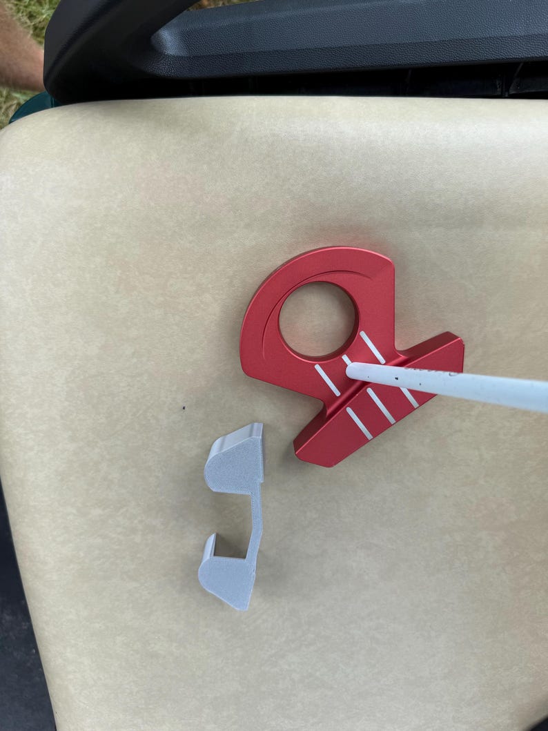 May include: A red golf putter with white stripes and a white shaft, along with a detached gray piece, rests on a beige surface. The putter has a circular opening and a unique design.